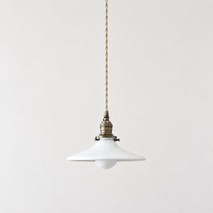 May include: A pendant light fixture featuring a glossy white ceramic shade with a wide, shallow profile. The lamp is suspended by a twisted beige fabric cord and includes a metallic brass-toned socket assembly with visible side screws.