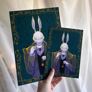 Könnte beinhalten: Zwei Kunstdrucke mit einer kaninchenartigen Figur im Anzug, die eine Tasse Tee hält. Die Drucke haben einen dunkelblauen Hintergrund mit einem goldenen, verzierten Rand. Das Kaninchen hat gelbe Augen und trägt eine Taschenuhr.