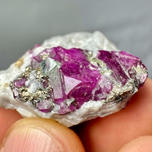 Könnte beinhalten: Nahaufnahme einer natürlichen Mineralprobe mit leuchtend tiefrosa Kristallen, die in eine weiße Matrix eingebettet sind. Die Kristalle haben eine prismatische Form, und die Probe weist stellenweise einen metallischen Glanz auf. Das Mineral wird in einer Hand gehalten.