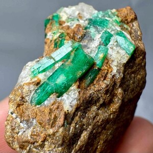 May include: A rough-cut emerald specimen featuring vibrant green crystal formations embedded in a matrix of brown and white rock. The emeralds have a crystalline structure, and the rock has a rough, textured surface.