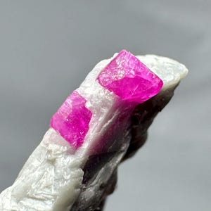 Puede incluir: Primer plano de un espécimen mineral con vibrantes cristales de color rosa intenso. Los cristales están bien definidos y se asientan sobre una matriz de piedra blanca y gris. La iluminación resalta la estructura cristalina y el color del mineral.