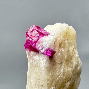 May include: A close-up of a raw mineral specimen. A cluster of bright pink crystals sits on top of a rough, off-white stone. The crystals have a faceted appearance, and the stone has a textured surface. The background is a plain grey.