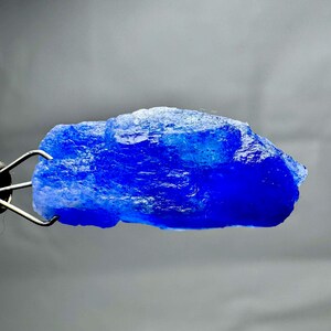 May include: A vibrant, raw blue gemstone held by metal tweezers. The gemstone has a crystalline structure and a deep, saturated blue color. The rough texture and irregular shape suggest it is in its natural, unpolished state.