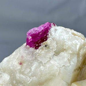 Pode incluir: Uma imagem em close-up de um cristal de rubi bruto e não cortado. O rubi é de uma cor magenta vibrante e está sobre uma matriz de rocha cristalina branca. A rocha tem uma superfície áspera e texturizada, e o fundo é cinza claro.