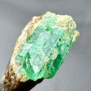 Könnte beinhalten: Ein leuchtend grüner Smaragdkristall mit rechteckiger Form, eingebettet in eine Matrix aus beigem und braunem Gestein. Die Oberfläche des Smaragds reflektiert das Licht und zeigt seine natürlichen Facetten und seine kristalline Struktur. Eine Nahaufnahme zeigt die Klarheit des Edelsteins.