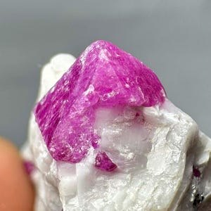 Pode incluir: Close-up de uma formação de cristais de rubi bruto. O cristal principal é um vermelho-rosado vibrante, com formato piramidal. Ele está situado em um fundo de rocha branca e texturizada. Pequenos fragmentos de rubi também são visíveis.