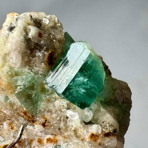 Könnte beinhalten: Nahaufnahme einer leuchtend grünen Mineralkristallformation. Der Kristall hat eine auffällige smaragdgrüne Farbe, eine rechteckige Form und sichtbare Facetten. Er ist vor dem Hintergrund einer hellen, strukturierten Matrix angeordnet.