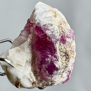 Puede incluir: Un espécimen de cristal de rubí en bruto con una mezcla de tonos blancos, rosas y dorados. La gema en bruto tiene una estructura cristalina y está sujeta por una herramienta de metal. El color rosa intenso del rubí contrasta con la matriz blanca.