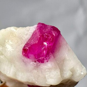 Puede incluir: Una vibrante formación de cristal rosa intenso sobre una base mineral blanca y texturizada. El cristal está bien definido con múltiples facetas, reflejando la luz. La composición general muestra un espécimen de gema natural y en bruto.