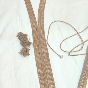 Pode incluir: Guarnição e cordão trançados dourados, provavelmente para embelezar roupas ou artesanato. A guarnição tem formato em V, com um pequeno grupo de elementos decorativos e um pedaço de cordão. Em uma superfície branca.