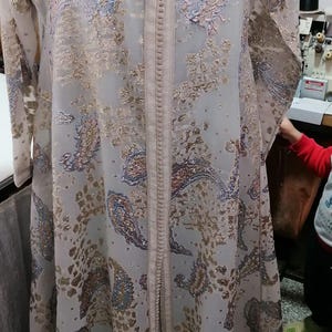 Op de afbeelding: Een lichtgrijze jurk met lange mouwen en gouden en blauwe paisley patronen. Het kledingstuk heeft een V-hals en een centrale lijn met decoratieve stiksels. De stof lijkt licht en vloeiend, ideaal voor warmer weer. De jurk is waarschijnlijk gemaakt van een delicaat materiaal.