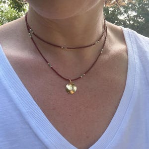 Puede incluir: Un collar en capas con un hilo de cuentas burdeos y un colgante dorado. El collar se lleva sobre una camiseta blanca con cuello en V. El colgante es un pequeño disco redondo de oro.