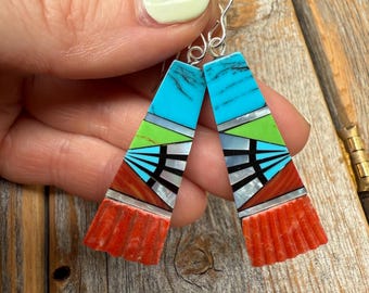 May include: A pair of colourful, geometric earrings. The earrings feature a turquoise top, green and silver middle, and a red, textured bottom. The design includes black and white accents. The earrings are held up by silver hooks.