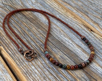 May include: A brown leather cord necklace with a strand of multi-colored gemstone beads. The beads are a mix of red, brown, green, and yellow.