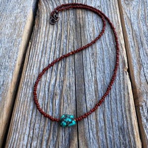 May include: A handcrafted necklace with a central turquoise stone with dark brown matrix, strung on a delicate strand of small, reddish-brown beads. The necklace has a copper-coloured clasp and is displayed on a rustic wooden surface.