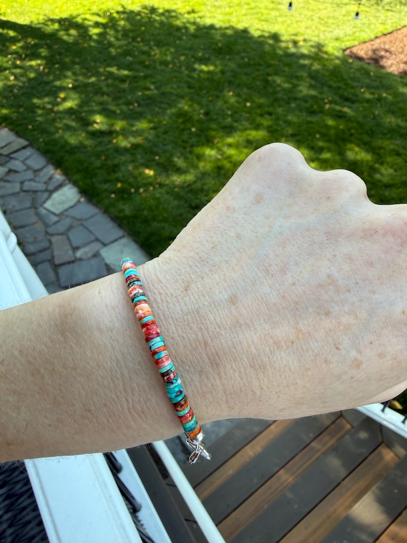 May include: A colorful beaded bracelet featuring turquoise, orange, and red disc-shaped beads. The bracelet is strung on a flexible cord and has a silver-colored clasp. The beads are approximately 0.25 inches in diameter.