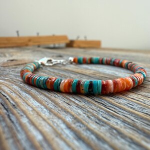 May include: A colorful beaded bracelet with turquoise, orange, red, and brown beads. The bracelet is strung on a silver chain and clasp. The beads are flat and round, creating a vibrant and eye-catching accessory.