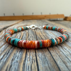 May include: A colorful beaded bracelet featuring a variety of orange, turquoise, and white disc-shaped beads. The bracelet is strung on a flexible cord and secured with a silver clasp. The bracelet is displayed on a wooden surface.