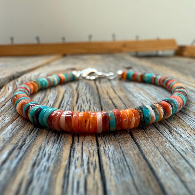 May include: A colorful beaded bracelet featuring a variety of orange, turquoise, and white disc-shaped beads. The bracelet is strung on a flexible cord and secured with a silver clasp. The bracelet is displayed on a wooden surface.