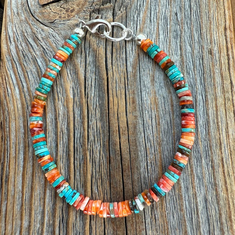 May include: A colorful beaded bracelet with turquoise, orange, and brown disc-shaped beads. The bracelet has a silver clasp and is displayed on a wooden surface. The beads are strung together to form a flexible band.