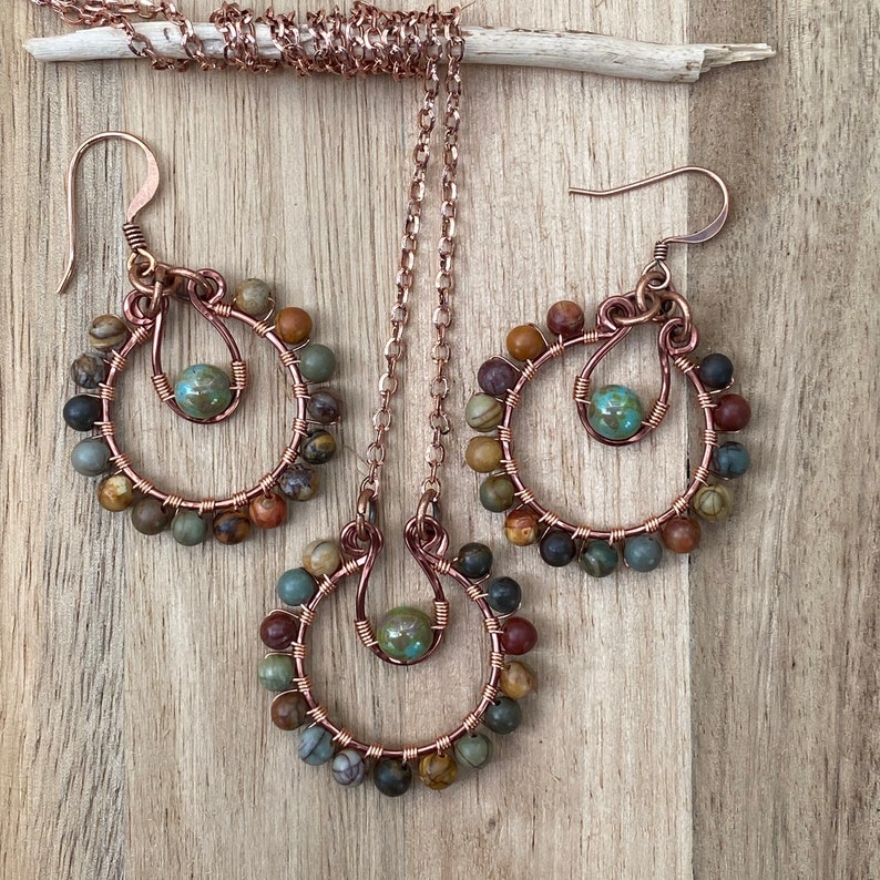 May include: A copper jewelry set featuring a necklace and matching earrings. Each piece has a circular design with a central turquoise bead and is adorned with a variety of earth-toned beads. The set is displayed on a wooden surface.