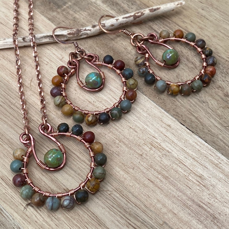 May include: A copper-toned jewelry set featuring a necklace and earrings. The necklace has a pendant with a green bead, and the earrings are hoop-shaped. Each piece is adorned with small, multi-colored beads, creating a natural, earthy aesthetic.
