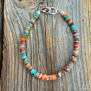 May include: A colorful beaded bracelet with turquoise, orange, and red-brown beads. The beads are small and round, strung together with a silver clasp. The bracelet is displayed on a weathered wooden surface.