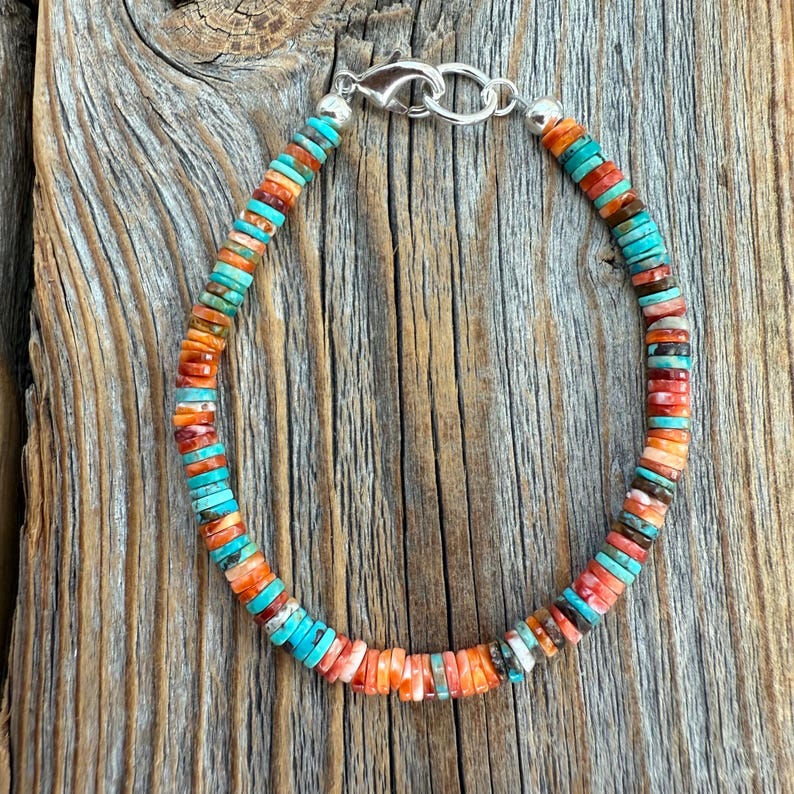 May include: A colorful beaded bracelet with turquoise, orange, and red-brown beads. The beads are small and round, strung together with a silver clasp. The bracelet is displayed on a weathered wooden surface.