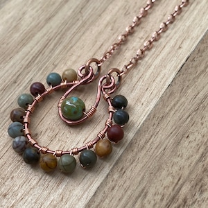 May include: A handcrafted copper wire necklace with a pendant featuring a central green bead and two U-shaped designs adorned with small, colorful beads. The necklace chain is also copper-colored, creating a cohesive and earthy aesthetic.