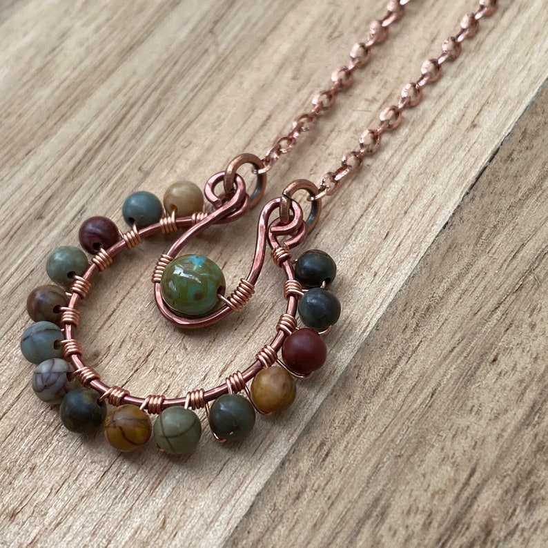 May include: A handcrafted copper wire necklace with a pendant featuring a central green bead and two U-shaped designs adorned with small, colorful beads. The necklace chain is also copper-colored, creating a cohesive and earthy aesthetic.