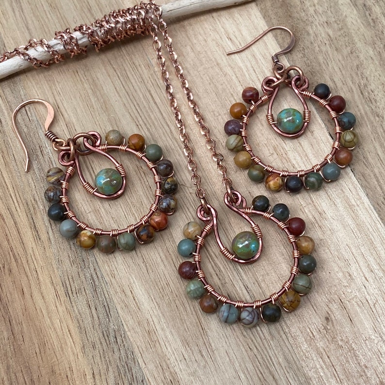 May include: A handcrafted jewelry set featuring copper wire earrings and a necklace. The earrings and pendant have a circular design, adorned with small, multi-colored beads and a central turquoise-colored bead. The set is displayed on a wooden surface.