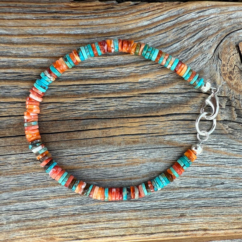 May include: A colorful beaded bracelet featuring turquoise, orange, and red disc-shaped beads. The bracelet has a silver clasp and is displayed on a wooden surface. The beads are strung together to form a flexible, circular design.