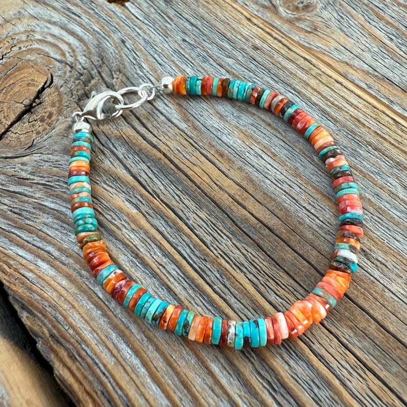 May include: A colorful beaded bracelet with turquoise, orange, and red-brown beads. The bracelet is strung on a silver chain with a lobster clasp. The beads are small and round, and the bracelet is displayed on a wooden surface.