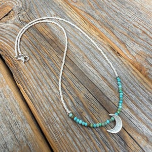 May include: A silver crescent moon pendant necklace with turquoise and white beaded accents. The necklace features a delicate white beaded chain and a silver clasp. The necklace is displayed on a rustic wooden surface.