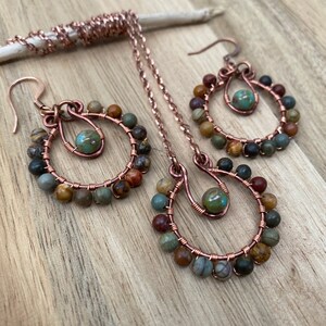 May include: A copper jewelry set featuring a necklace and earrings. The earrings and pendant have a circular design with multi-colored beads and a turquoise-colored bead in the center. The jewelry is displayed on a wooden surface.