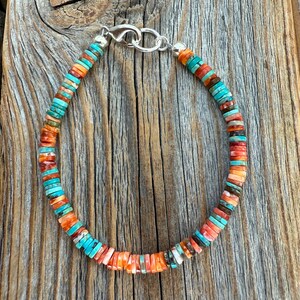 May include: A colorful beaded bracelet featuring turquoise, orange, and red-toned beads. The bracelet is strung on a silver chain with a clasp. The beads are small and round, creating a vibrant and eye-catching accessory. The bracelet is displayed on a wooden surface.