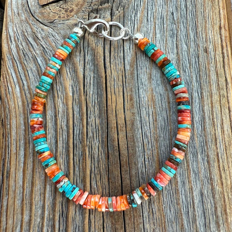 May include: A colorful beaded bracelet featuring turquoise, orange, and red-toned beads. The bracelet is strung on a silver chain with a clasp. The beads are small and round, creating a vibrant and eye-catching accessory. The bracelet is displayed on a wooden surface.