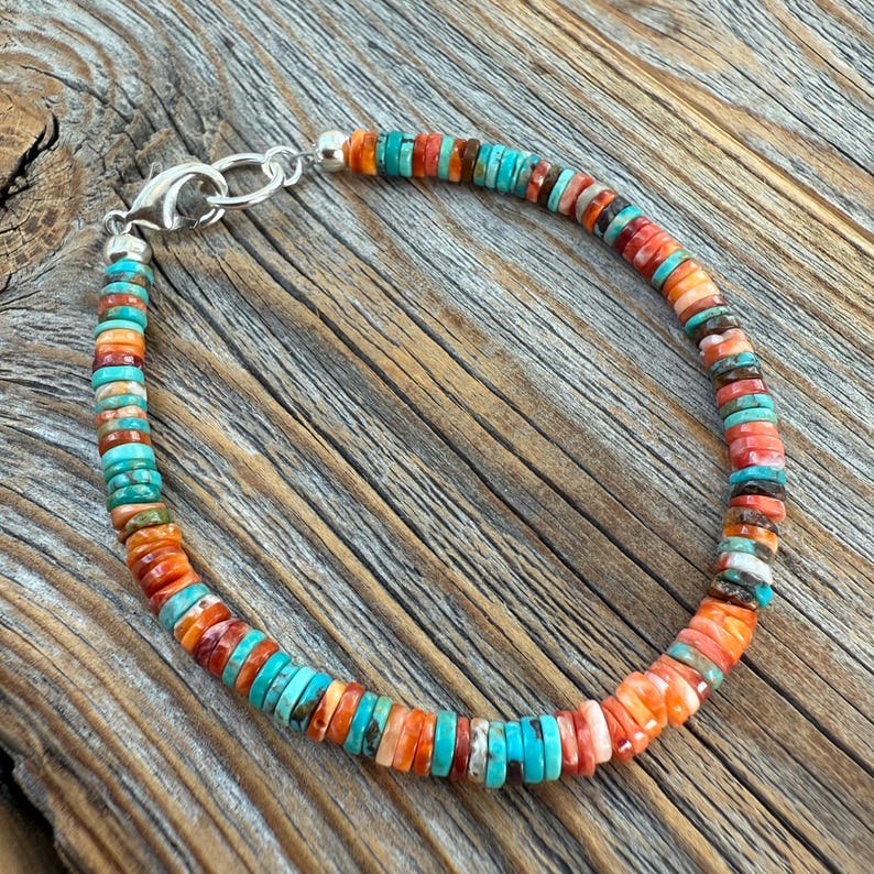 May include: A colorful beaded bracelet with a silver clasp. The bracelet features small, flat beads in shades of turquoise, orange, red, and brown. The beads are strung together to form a circular shape, perfect for wrist wear.