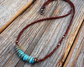 May include: A turquoise and copper beaded necklace with a brown cord. The necklace is on a wooden surface.