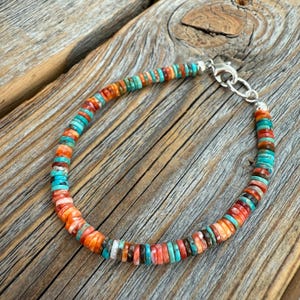 May include: A colorful bracelet featuring small, flat, round beads in shades of orange, turquoise, red, and brown. The beads are strung together with a silver clasp. The bracelet is displayed on a wooden surface.