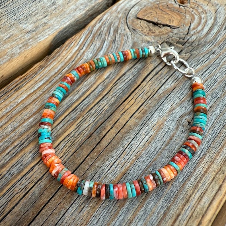 May include: A colorful bracelet featuring small, flat, round beads in shades of orange, turquoise, red, and brown. The beads are strung together with a silver clasp. The bracelet is displayed on a wooden surface.