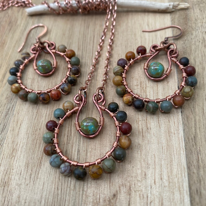 May include: A copper jewelry set featuring a necklace and a pair of earrings. Each piece has a circular design with a central turquoise bead and is adorned with small, multi-colored beads. The set is displayed on a wooden surface.