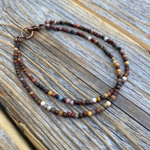 May include: A double strand necklace made with small, multi-colored gemstone beads. The beads are a mix of brown, red, yellow, white, and gray. The necklace has a copper clasp.