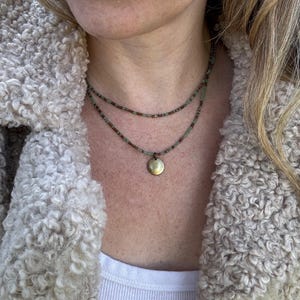 以下が含まれることがあります： 小さな丸い金のペンダントが付いた、重ね付けのネックレス。ネックレスは、小さな緑と茶色のビーズで作られています。