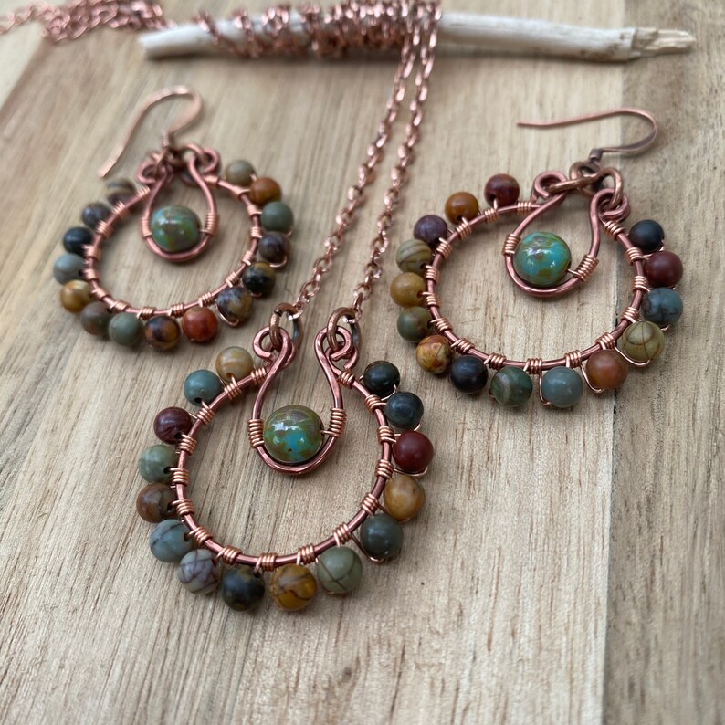 May include: A handcrafted jewelry set featuring a necklace and earrings. The set includes a copper wire pendant and earrings, each adorned with colorful beads and a central turquoise-colored bead. The jewelry is displayed on a wooden surface.