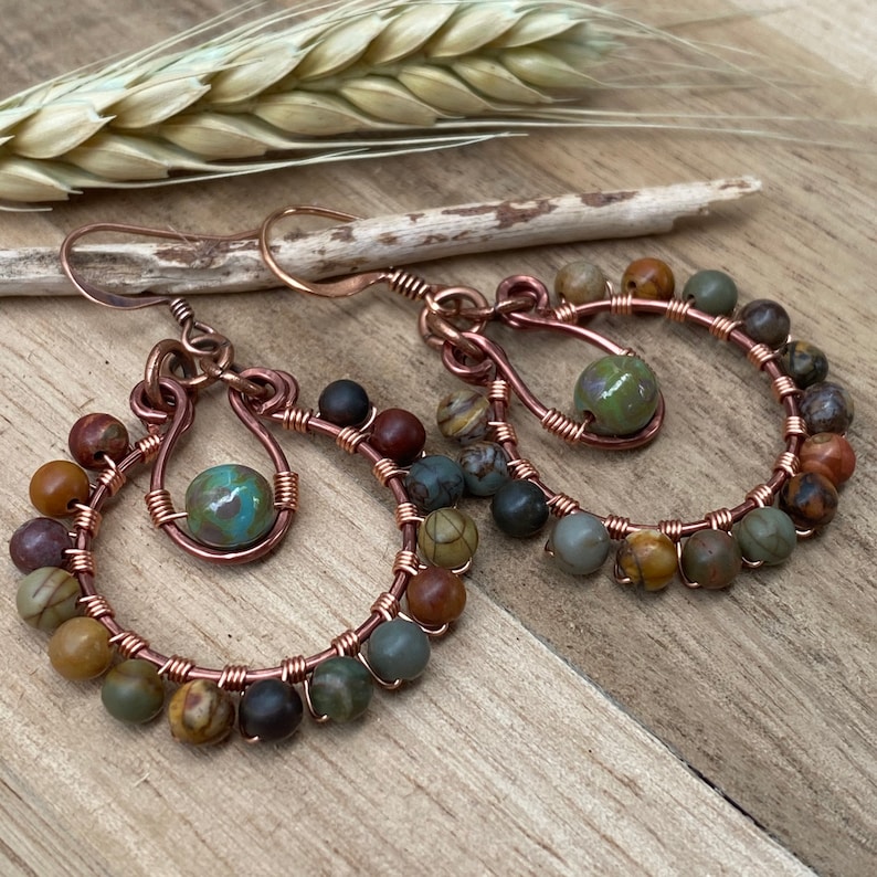 May include: Handmade copper wire hoop earrings with colorful gemstone beads. The earrings feature a circular design with a central teardrop shape and are adorned with various earth-toned beads, including green, brown, and black. The earrings are displayed on a wooden surface.