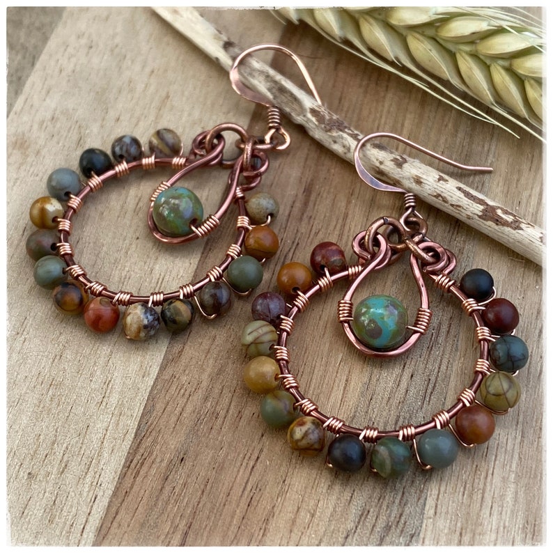 May include: A pair of handcrafted copper wire hoop earrings. Each earring features a circular design adorned with a variety of colorful, round beads in shades of brown, green, and black. A single turquoise bead hangs in the center of each earring.