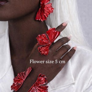 May include: Red flower-shaped jewelry set, including earrings, a ring, and a bracelet. The flower size is 5 cm. The jewelry has a metallic finish and is worn on a person with dark skin and white hair.