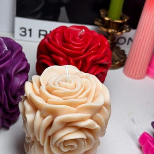 May include: Three decorative rose-shaped candles. One candle is red, one is purple, and one is a light peach colour.