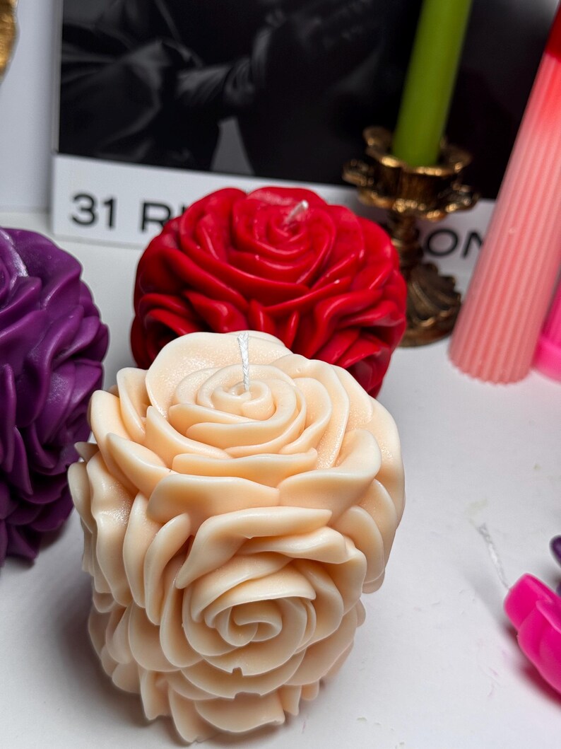 May include: Three decorative rose-shaped candles. One candle is red, one is purple, and one is a light peach colour.